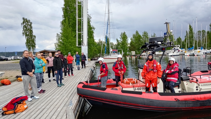 Kuvassa vasemmalla purjehtijakurssin oppilaita seisoo laiturilla, oikealla on Jyväskylän Järvipelastajien punainen ribbi-vene ja neljän hengen miehistö punaisissa pelastuspuvuissa